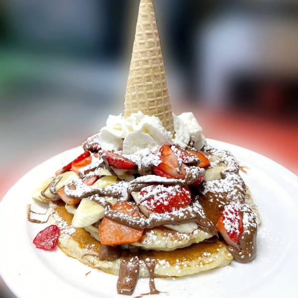 Stack of pancakes with strawberries, bananas, chocolate, whipped cream, and a waffle cone.
